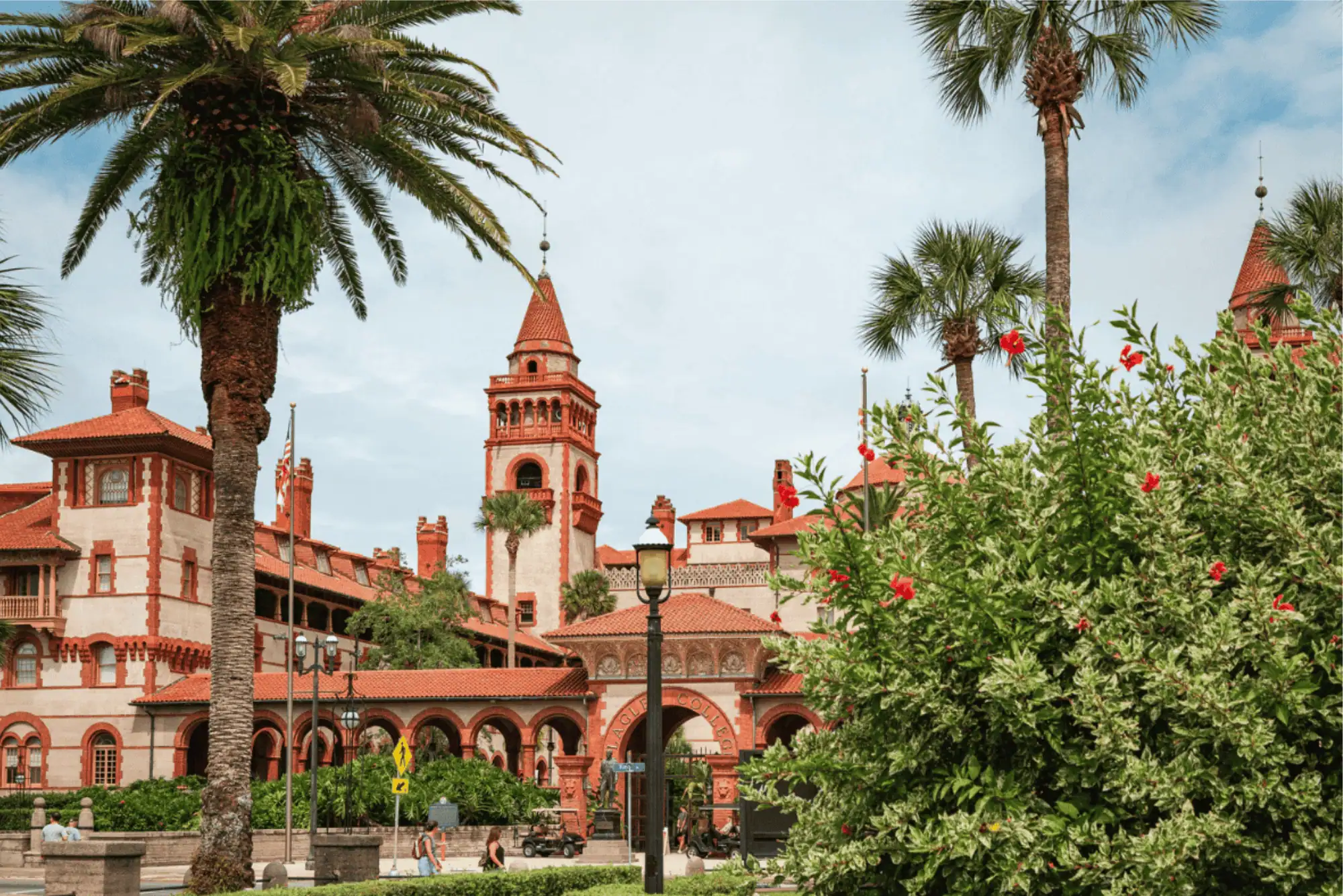 The historic Spanish Renaissance-style architecture of Flagler College in St. Augustine, featuring its iconic terracotta-roofed towers, arched walkways, tall palm trees, and blooming red hibiscus bushes in the foreground