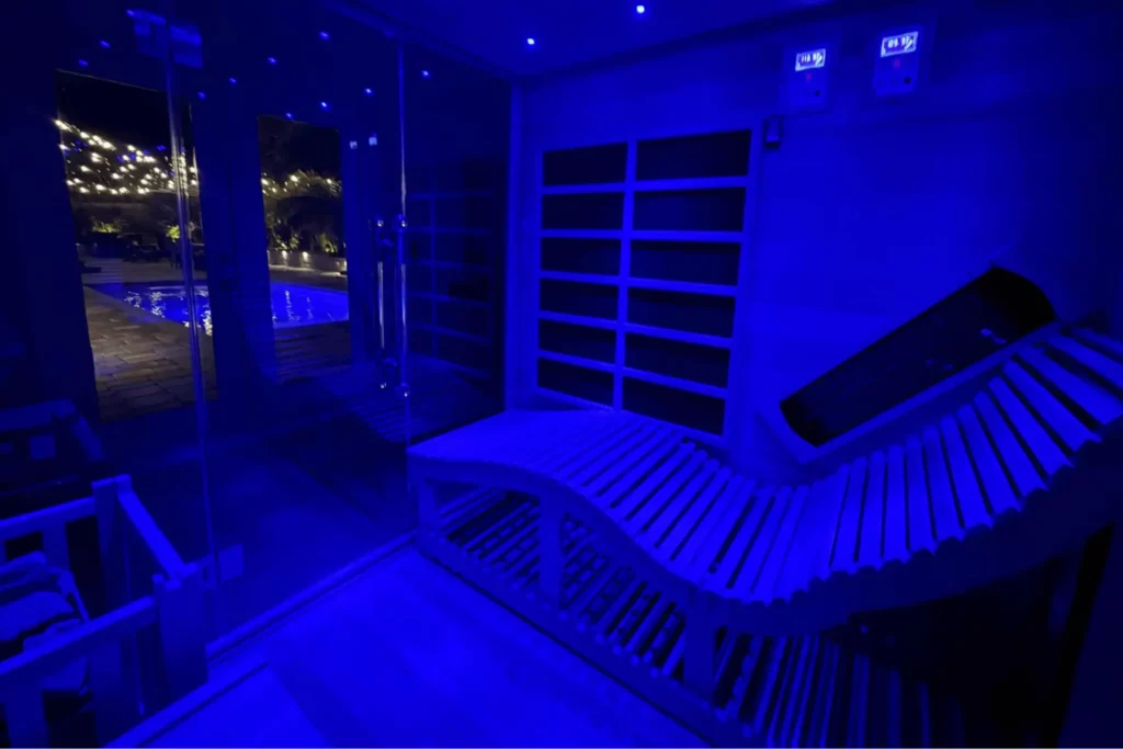 Interior view of a modern indoor sauna illuminated by deep blue LED lighting, featuring an ergonomic curved wooden lounger, infrared panels, and a glass wall overlooking an outdoor pool at night