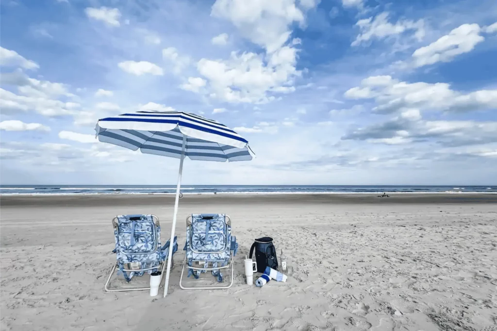 A blue and white striped beach umbrella over two patterned beach chairs on a wide sandy shore under a cloudy blue sky