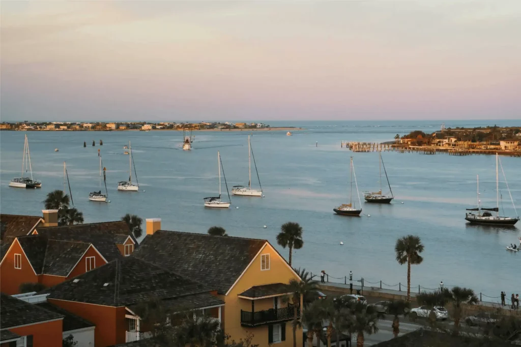 Elevated view of a coastal inlet at dusk in St. Augustine, featuring sailboats anchored in calm blue water, rooftops of colorful houses with palm trees in the foreground, and Vilano Beach across the bay