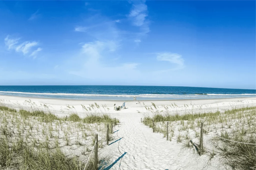 Wide panoramic view of a sandy beach path lined with wooden rope posts leading through sea grass dunes onto the wide sandy shore and deep blue ocean at Vilano Beach under a bright blue sky