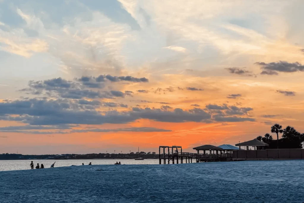 A vibrant golden orange sunset over the water at Vilano Beach, with silhouetted beachgoers, a wooden pier, and covered pavilions along the white sand shore