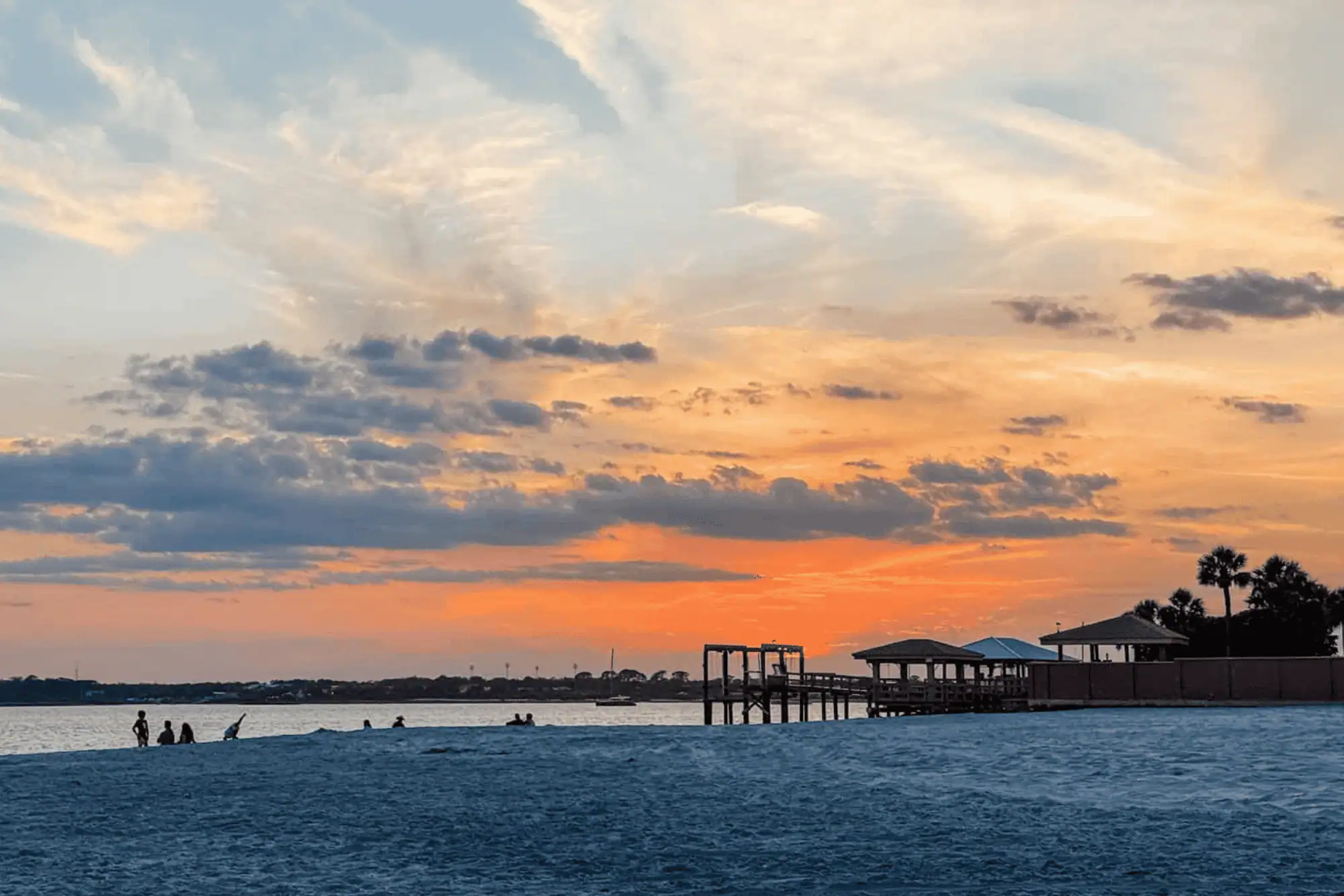 A vibrant golden orange sunset over the water at Vilano Beach, with silhouetted beachgoers, a wooden pier, and covered pavilions along the white sand shore