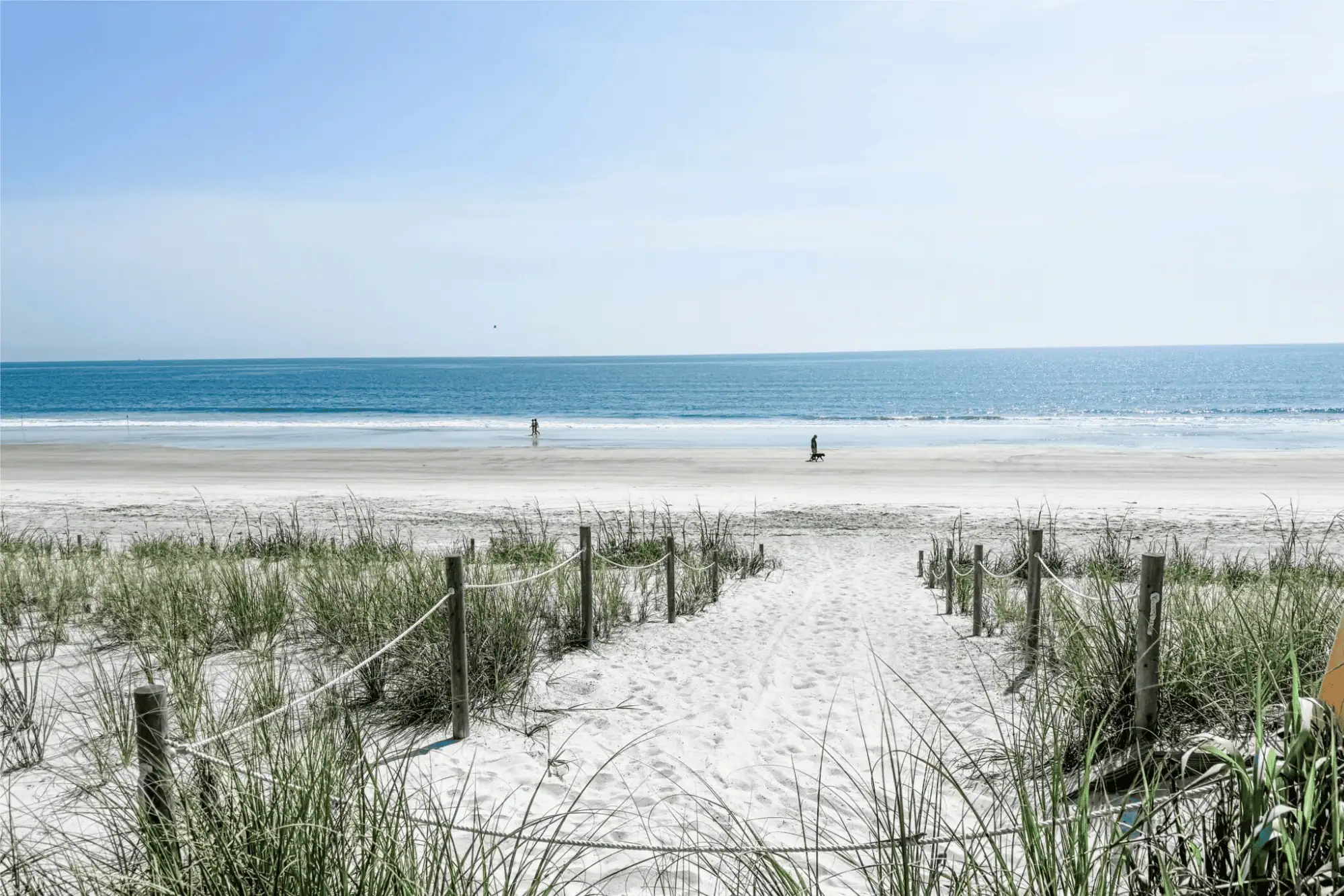 A sandy path lined with wooden posts and rope railings leading through sea grass toward the wide sandy shore and blue waters of Vilano Beach