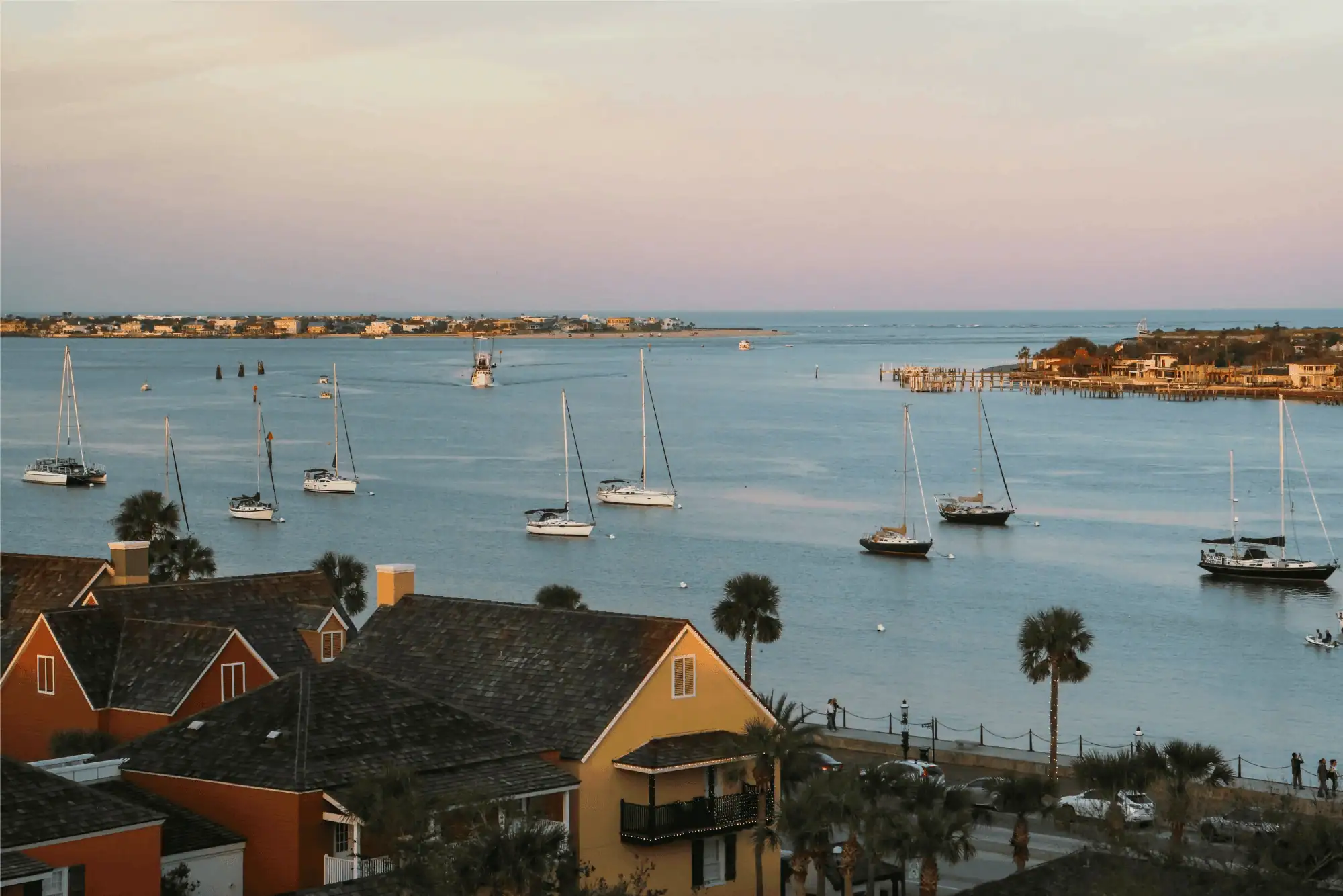 Elevated view of a coastal inlet at dusk in St. Augustine, featuring sailboats anchored in calm blue water, rooftops of colorful houses with palm trees in the foreground, and Vilano Beach across the bay