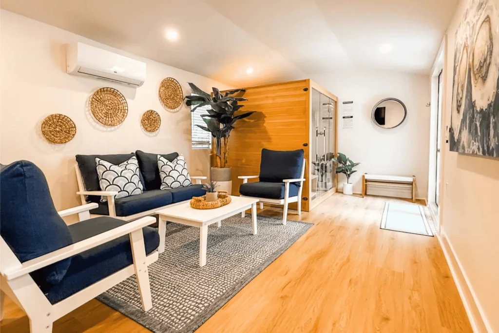 A bright wellness room featuring a navy blue sofa and armchairs, a white coffee table on a patterned rug, woven wall decor, and a wooden indoor sauna with a glass door