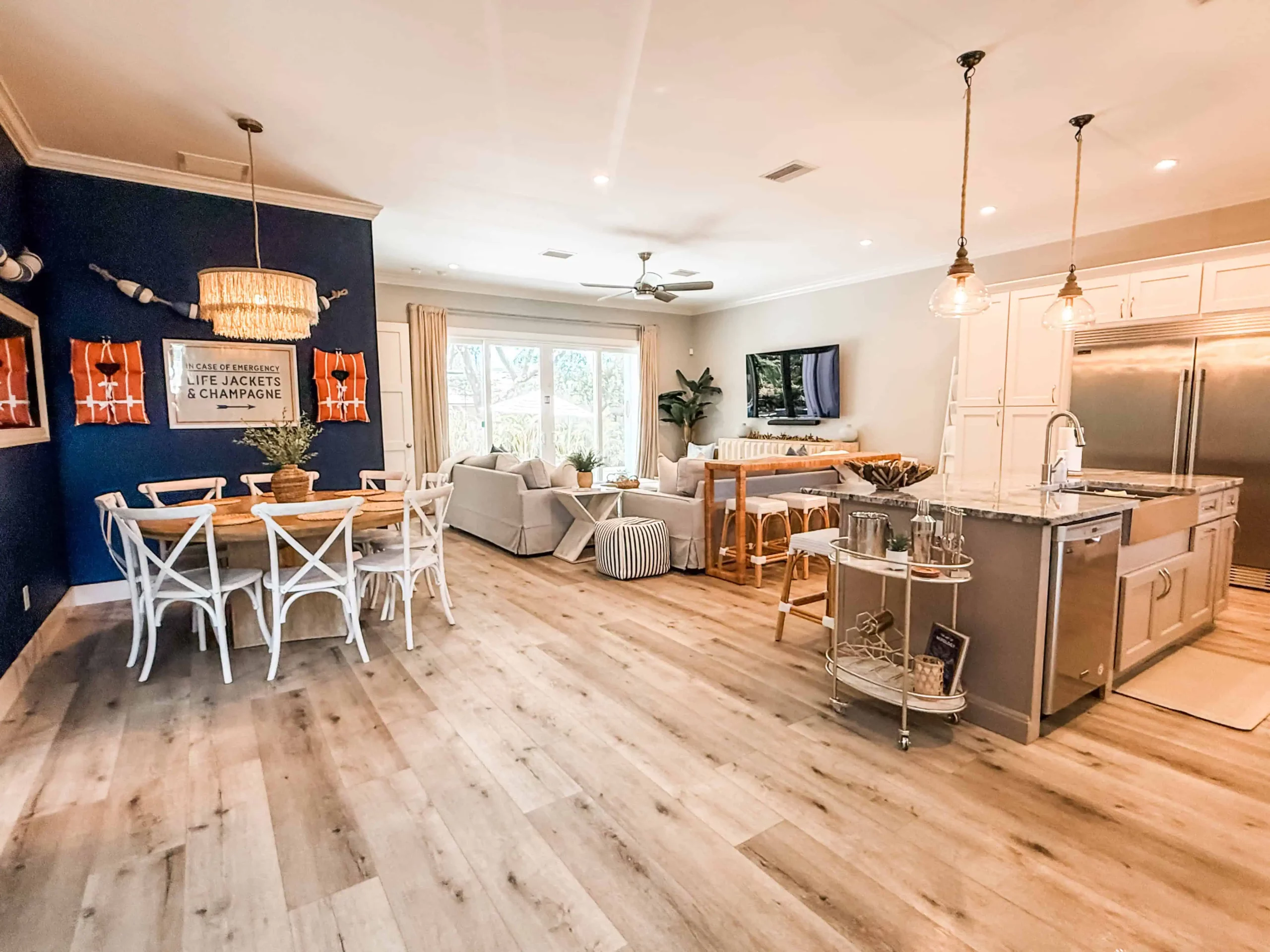 Bright open-concept living space featuring a coastal dining area with a navy blue accent wall, a modern kitchen island, and a light-filled living room with light wood floors.